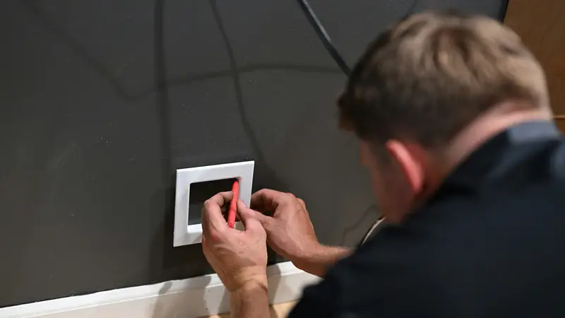 Elektriker setzt einen Schalterrahmen an einer dunkelgrauen Wand und zeichnet die Position an