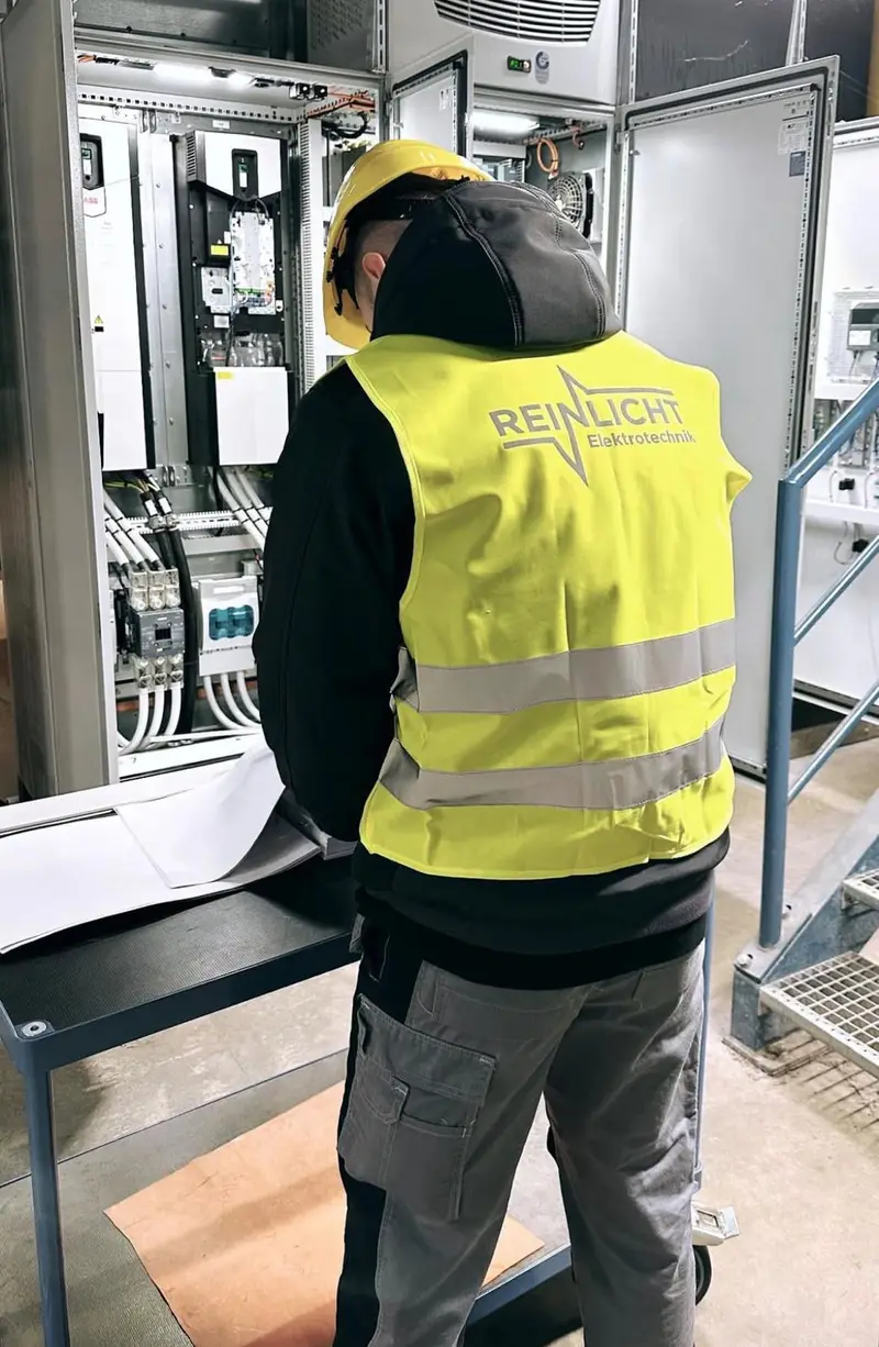Techniker von Reinlicht Elektrotechnik mit Schutzhelm und Warnweste prüft Pläne vor einem offenen Schaltschrank