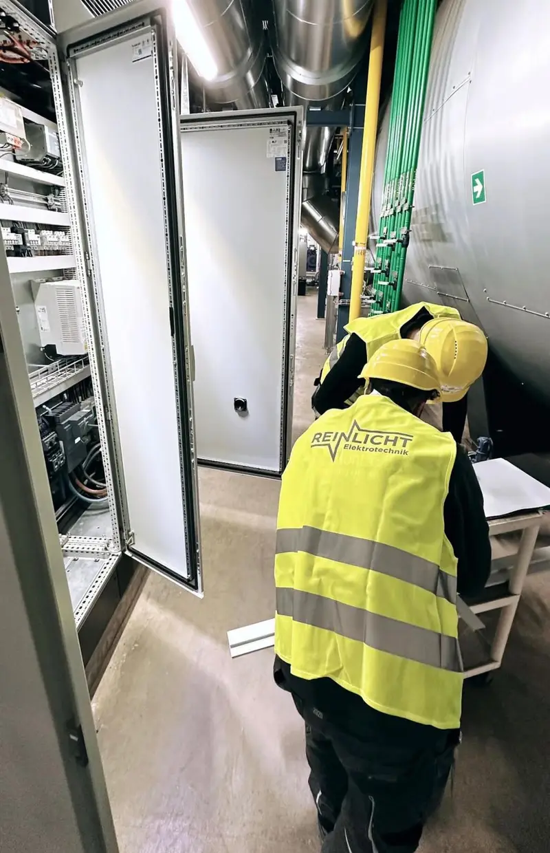 Zwei Techniker von Reinlicht Elektrotechnik mit Schutzhelmen an offenen Schaltschränken in einer Technikzentrale