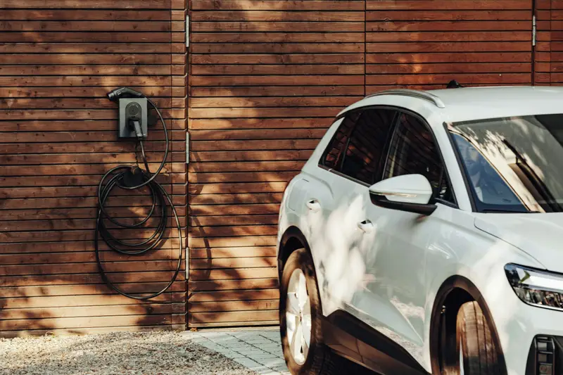 Wallbox mit aufgewickeltem Ladekabel an einer Holzfassade, daneben ein weißes Auto
